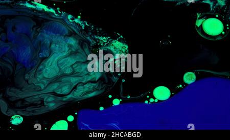 Close up of green and blue color oil paint being added into black inks. Mixed liquid paints texture on black background, art and creativity concept. Stock Photo