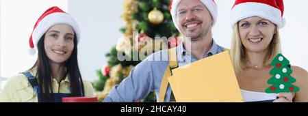 Group of couriers in santa claus hats standing near christmas tree and holding many gift boxes Stock Photo