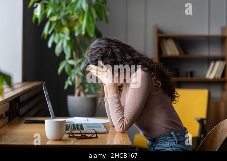 Exhausted unhappy woman sit at laptop hide face in hands tired work overtime suffer from burnout Stock Photo