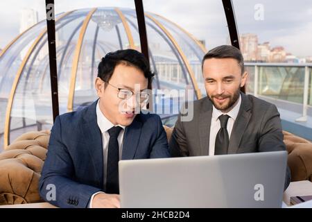 Two successful businessmen in suits looking at laptop display and discussing data while having meeting in cafe Stock Photo