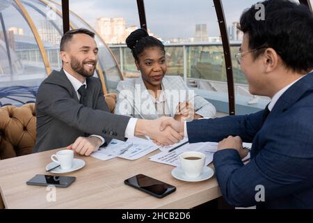 Two intercultural successful businessmen shaking hands over papers on table after negotiation about signing new contract Stock Photo