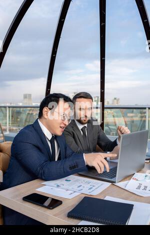 Young confident Asian businessman explaining his Caucasian colleague online data at working meeting in cafe Stock Photo