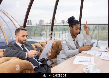 Two intercultural coworkers in formalwear scrolling in mobile phones while sitting on sofa by table in cafe Stock Photo
