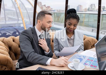 Two intercultural colleagues looking at financial paper while discussing it by table at working meeting in cafe Stock Photo