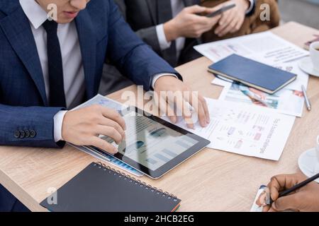 Contemporary economist in suit looking through charts and diagrams in tablet while sitting by table among coworkers Stock Photo
