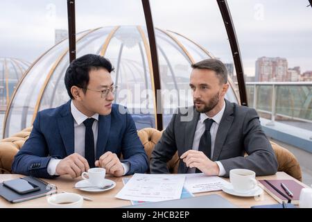Two young intercultural business partners having tea and planning work while having meeting by table in cafe Stock Photo