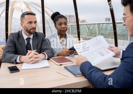 Two intercultural colleagues looking at Asian businessman with financial papers during discussion of data at working meeting Stock Photo