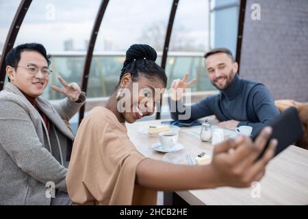 Young cheerful businesswoman making selfie with two male colleagues by table served with drinks and plates with desserts Stock Photo
