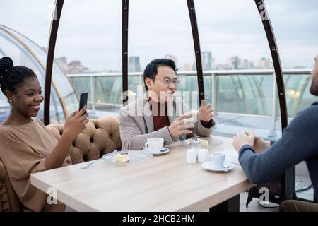 Happy young African woman taking photo of her colleague while sitting by table and having tea with cheesecake Stock Photo
