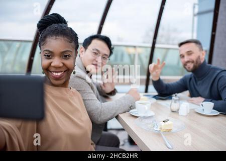 Three happy intercultural colleagues looking in smartphone camera while pretty African woman making selfie with two men Stock Photo