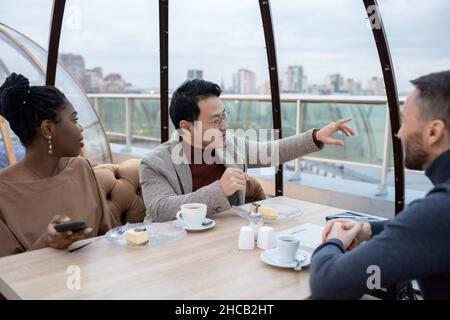 Contemporary young colleagues having discussion by dessert while one of them pointing at something curious behind window Stock Photo