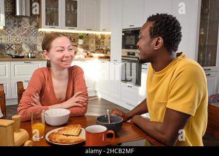 Happy young intercultural couple chatting by breakfast in the morning while sitting by wooden kitchen table Stock Photo