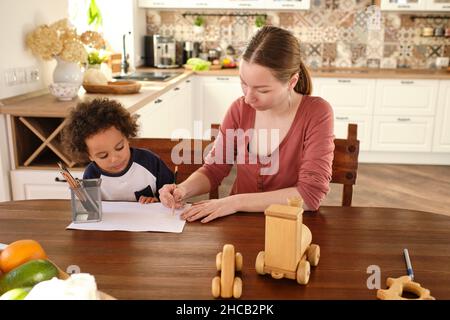 Young woman drawing something with pencil on sheet of paper while teaching her little son in the kitchen Stock Photo