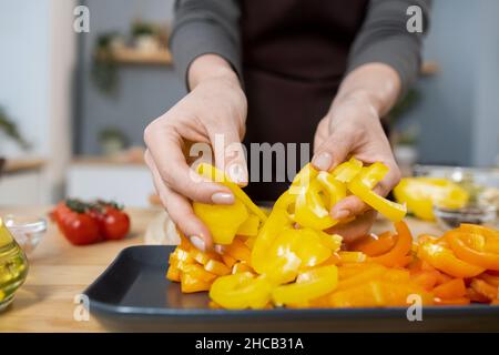 Hands of female putting chopped fresh yellow capsicum on tray while cooking vegetarian stew for dinner in the kitchen Stock Photo