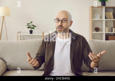 Webcam view of man in glasses sitting on couch at home, looking at camera and talking Stock Photo