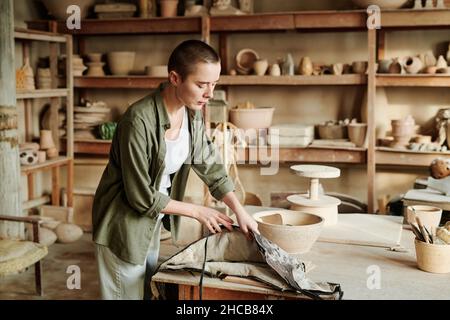 Craftswoman standing at her workplace and taking her apron, she preparing for her work in workshop Stock Photo