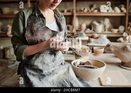 Close-up of woman in apron working online on her mobile phone while working with clay in the workshop Stock Photo
