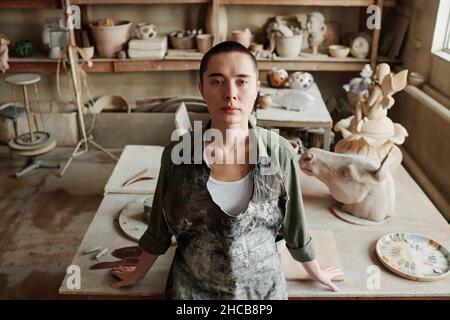 Portrait of young craftswoman in apron looking at camera standing at her workplace creating ceramic vases in the workshop Stock Photo