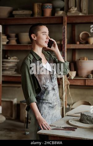 Young craftsperson in apron having a conversation on mobile phone while standing in the workshop Stock Photo