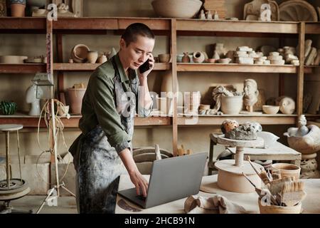 Young sculptor working on her laptop during conversation on mobile phone in the workshop Stock Photo