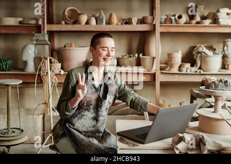 Young woman gesturing and smiling while talking online on laptop during her work in ceramic studio Stock Photo