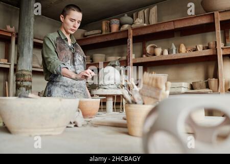 Young woman using stick to make little details on the clay sculpture while working in the studio Stock Photo