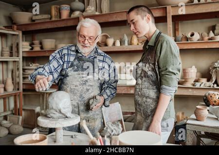 Senior man in apron holding the lesson for woman in pottery workshop, he teaching her to make sculpture from clay Stock Photo