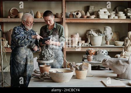 Senior sculptor pointing at tablet pc and showing his assistant the ceramic sculpture during their work in pottery studio Stock Photo