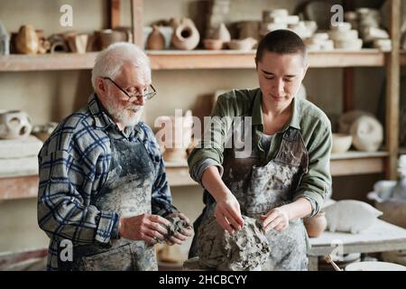 Young woman learning to make sculpture from clay with her teacher standing near by and teaching her in the workshop Stock Photo