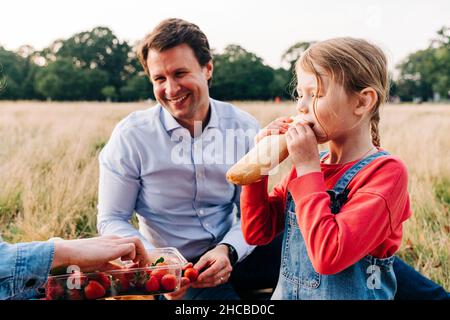 Girl eating loaf of bread at picnic with family in park Stock Photo