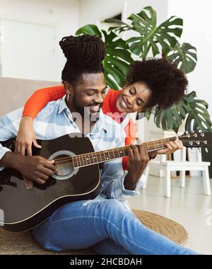 Curly haired woman embracing boyfriend playing guitar at home Stock Photo