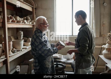 Senior potter discussing ceramic vases with his assistant while they standing in the pottery studio Stock Photo