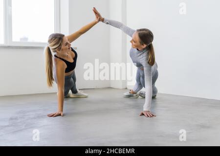 Young women practicing yoga together giving high five holding hands and standing in side plank pose on rubber mats at fitness studio Stock Photo