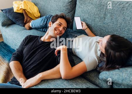 Happy young man looking at girlfriend with mobile phone on sofa at home Stock Photo
