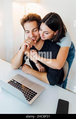 Young couple embracing while working on laptop at home Stock Photo