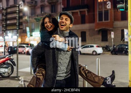 Cheerful boyfriend carrying girlfriend piggyback while walking in city Stock Photo