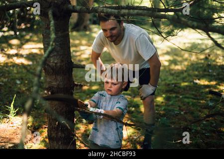 Father looking at son cutting branch of tree with hand saw Stock Photo