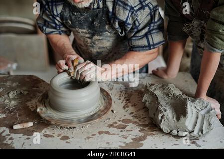 Close-up of pottery artist shaping clay on pottery wheel in workshop with his assistant Stock Photo