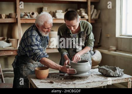 Young female potter making earthenware vase on pottery wheel with senior man helping her during their work in workshop Stock Photo
