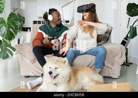 Smiling woman wearing virtual reality headset by man playing video game in front of dog at home Stock Photo