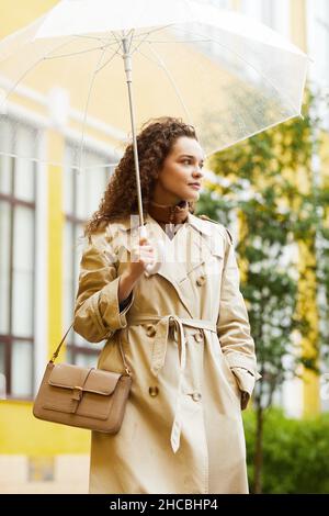 Vertical medium long portrait of young Caucasian woman wearing trench coat standing outdoors under umbrella on rainy day Stock Photo