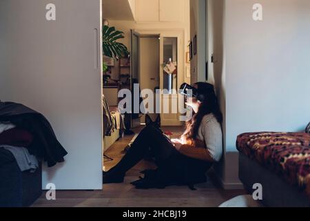 Woman with virtual reality headset sitting by wall at home Stock Photo