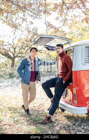 Young man sitting at van's trunk by friend in autumn forest Stock Photo