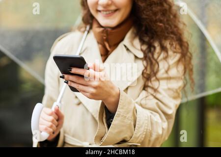 Horizontal medium close-up selective focus shot of Caucasian woman standing outdoors under transparent umbrella on rainy day using smartphone Stock Photo
