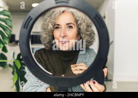 Senior woman applying foundation in front of ring light Stock Photo