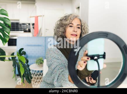 Senior woman applying foundation make-up in front of ring light at home Stock Photo