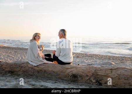 Couple talking to each other sitting on tree log at beach Stock Photo