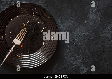 Dirty glossy black plate and a fork on a black textured background with copy space. Dirty and empty dishes after meals and holidays. A plate after a d Stock Photo