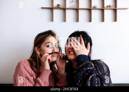 Smiling woman covering face with hand by friend making mustache from her hair Stock Photo