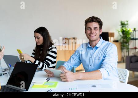 Smiling businessman with tablet PC sitting with colleague using smart phone at office Stock Photo